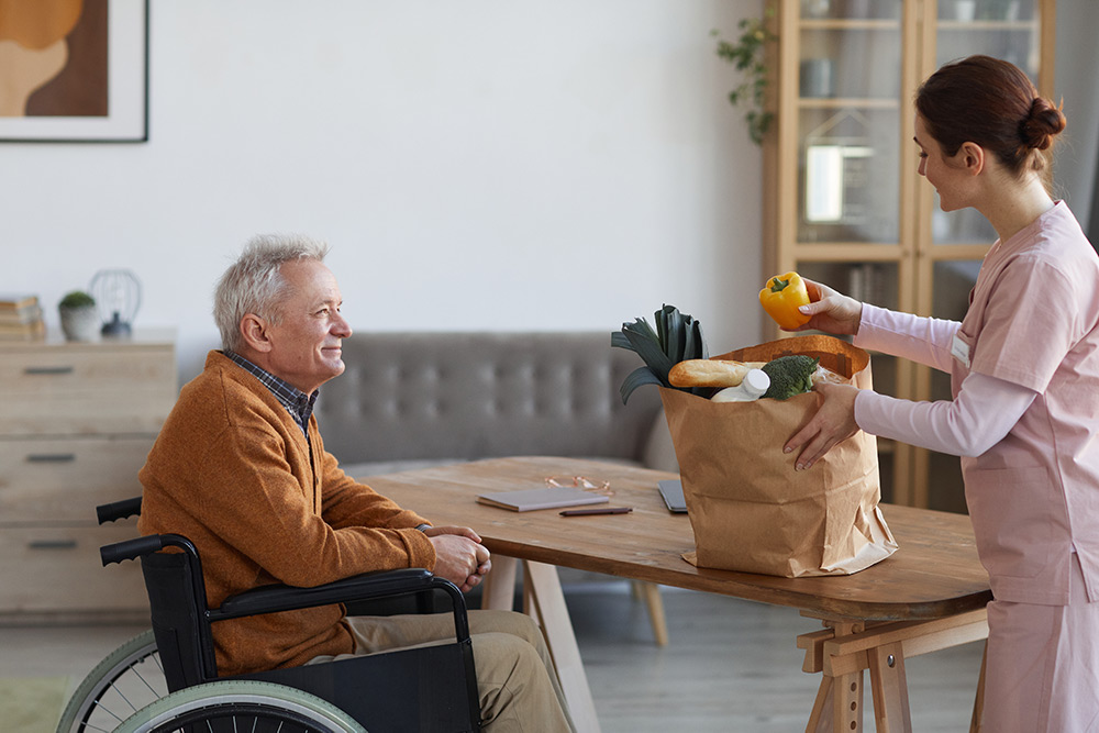 Carer unpacking groceries on table for senior man in wheelchair